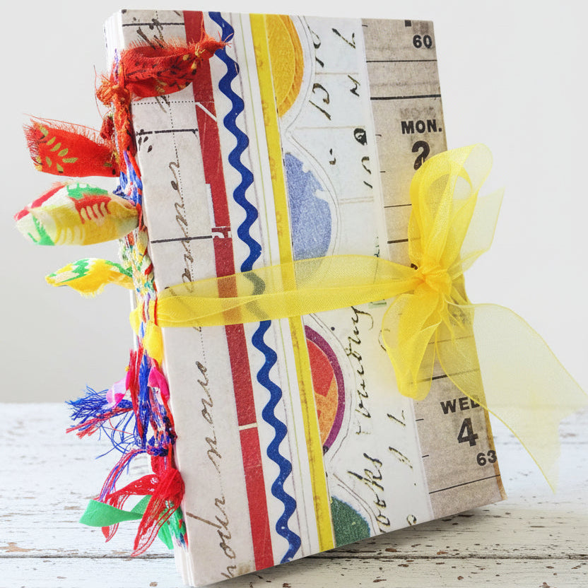 A handmade journal with a colorful soft cover, adorned with ribbons and decorative elements. The journal lies flat on a surface with its spine visible, showing the colorful embellishments.